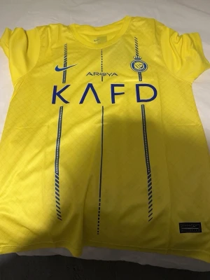 Al Nassr Ronaldo matchtröja gul - Gul Al Nassr matchtröja från Nike med blå detaljer, klubbmärke och sponsortryck framtill. På ryggen står det 'Ronaldo' och nummer 7 i blått. Tröjan har rund hals och är tillverkad i lätt, ventilerande material perfekt för fotbollsträning.