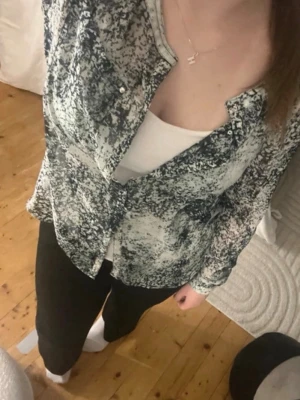 Mönstrad långärmad skjorta - Säljer en snygg långärmad skjorta med svartvitt (lite förg typ blå och grön aktigt) abstrakt mönster. Skjortan har knappar framtill och bröstfickor, perfekt för dig som gillar en unik och trendig stil. Materialet känns lätt och luftigt, vilket gör den skön att bära. Tunn och fin till sommaren med mera. Några knappar är borta längst ned men inget man tänker eller bryr sig om alls. Har bundle rabatt så kolla gärna in mer grejer! 