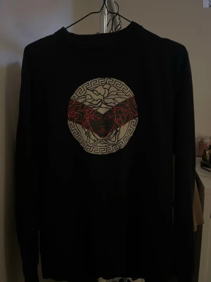 Svart långärmad tröja med Medusa-tryck - Svart långärmad tröja med stort Medusa-inspirerat tryck i vitt och rött på bröstet. Tröjan har rund halsringning och är gjord i ett mjukt material som passar perfekt till en streetwear-look. Snygg detalj med grafiskt mönster runt motivet.