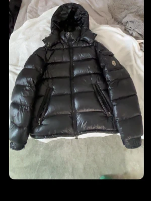 Svart dunjacka från Moncler - Snygg svart dunjacka från Moncler med glansig finish och klassisk puffer-design. Jackan har huva, två dragkedjeförsedda fickor framtill och ikonisk Moncler-logga på ärmen. Insidan har ett serietidningsmönster och innerficka. Perfekt för kalla vinterdagar.