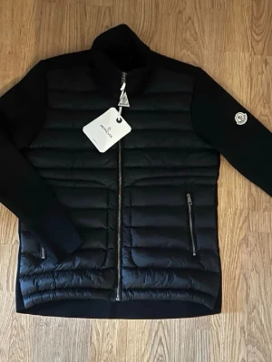 Svart dunjacka från Moncler - Snygg svart dunjacka från Moncler med quiltad front, två dragkedjeförsedda fickor och Moncler-logga på ärmen. Jackan har hög krage och raka ärmar i mjukare material, perfekt för kalla vinterdagar. Stängs med dragkedja framtill.