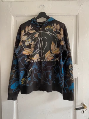 Ed Hardy Christian Audigier Hoodie - Säljer en svart hoodie från Christian Audigier med dragkedja, stora broderade blommor och döskallemotiv i gult, blått och beige. Hoodien har långa ärmar och detaljerade mönster både fram och bak. Christian Audigier är grundaren av Edhardy märket. Denna hoodie är under hans egna namn. Perfekt för dig som gillar statement-plagg!