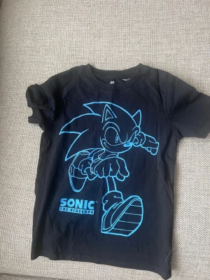 Svart Sonic the Hedgehog t-shirt H&M - Cool svart t-shirt från H&M med ett stort blått Sonic the Hedgehog-tryck på framsidan. Klassisk rund hals och korta ärmar. Perfekt för alla Sonic-fans som vill sticka ut med en snygg och bekväm t-shirt i bomull.( LITET HÅL SYNS SISTA BILDEN )
