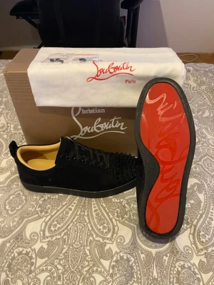 Svarta Louboutin sneakers med nitar - Snygga svarta sneakers från Christian Louboutin med klassisk röd sula och svarta spikar på tån. Skorna har lågt skaft, svarta skosnören och är tillverkade i mocka. Perfekta för dig som vill sticka ut med en lyxig och edgy stil. Medkommer även två små påsar med extra spikar samt, en dustbag