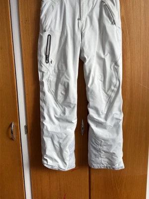 Vita täckbyxor från Peak Performance - Säljer ett par vita täckbyxor från Peak Performance med vattentäta dragkedjor och snygga sömdetaljer. Byxorna har flera fickor och är tillverkade i ett slitstarkt material som passar perfekt för vinterns alla äventyr.