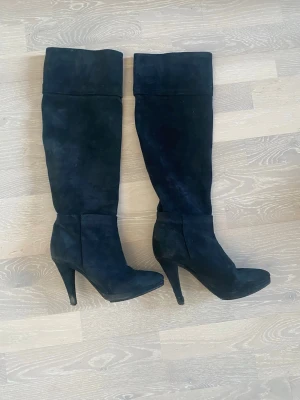 Svarta höga mockastövlar med klack - Snygga svarta stövlar i mocka med hög klack och spetsig tå. Stövlarna går upp över knäet och har en stilren, enkel design utan synliga detaljer eller dragkedjor. Perfekta för att lyfta vilken outfit som helst.