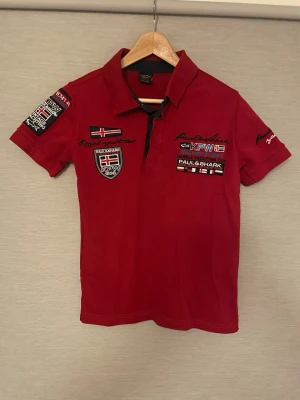 Röd pikétröja från Paul & Shark (Small) - Röd pikétröja från Paul & Shark. Tröjan har broderade märken och patchar på bröstet och ärmarna, samt klassisk krage och knäppning framtill. Snygga detaljer med flaggor och text, perfekt för en sportig look.