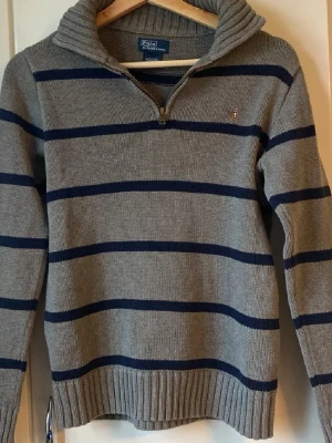 Gråblå randig stickad tröja Polo - Snygg stickad tröja från Polo Ralph Lauren i grått med mörkblå horisontella ränder. Tröjan har halv dragkedja, hög krage och ribbade muddar. Perfekt för dig som gillar klassisk och avslappnad stil.