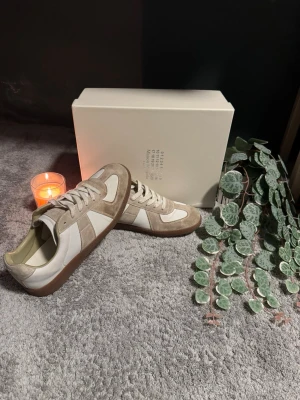 Maison Margiela Replica sneakers beige/vit - Snygga Maison Margiela Replica sneakers i beige mocka och vitt läder. Klassisk low-top modell med gummisula och vita skosnören. Perfekta för dig som gillar minimalistisk och stilren design med premiumkänsla.