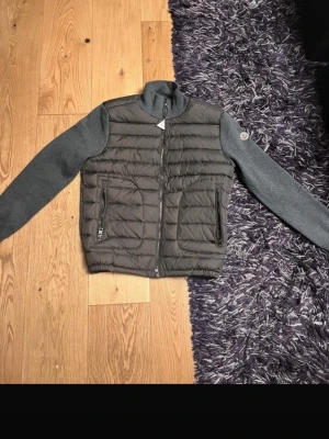 Moncler hybrid cardiganjacka grå/svart - Snygg cardiganjacka från Moncler med quiltad svart front och grå stickade ärmar. Jackan har två dragkedjefickor fram och hög krage. Moncler-logga på vänster ärm. Perfekt för lager-på-lager och streetwear-vibe.