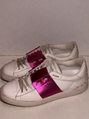 Valentino opens  - Snygga vita sneakers från Valentino med en metallicrosa bred rem över snörningen. Skorna har klassisk låg profil, vita snören och diskret logga på plösen. Tillverkade i läder med gummisula. Perfekta för dig som vill sticka ut med en färgklick.