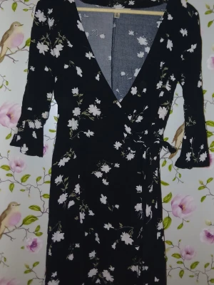 Svart blommig omlottklänning - Snygg svart kortklänning med vitt blommönster och omlottmodell. Klänningen har trekvartsärm och knytband i midjan för en smickrande passform. Perfekt för dig som gillar en feminin och trendig stil.