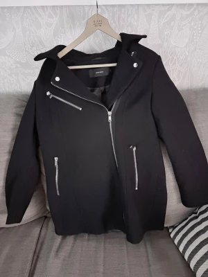 Svart rock från Vero Moda med dragkedjor - Snygg svart rock från Vero Moda med flera silvriga dragkedjor och en cool krage. Rocken har lång ärm och är tillverkad i ett tjockare material som passar perfekt till höst och vinter. Stängs med snedställd dragkedja framtill för en modern look.
