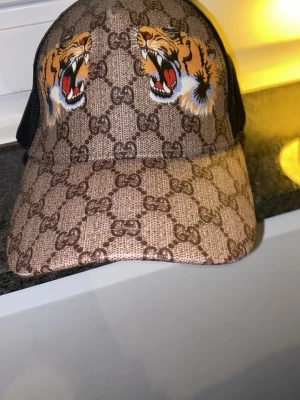 Gucci keps med tigertryck och GG-mönster - Säljer en brun Gucci keps med klassiskt GG-mönster och två stora tigerhuvuden framtill. Kepsen har svart mesh på sidorna och justerbar rem bak. Materialet är canvas framtill och mesh baktill, vilket ger en cool och trendig look.