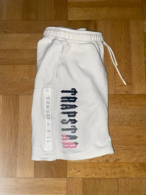 Vita Trapstar shorts med tryck - Säljer ett par vita shorts från Trapstar med färgglatt logotyptryck i svart, grått och rosa på vänster ben. Shortsen har resårmidja med dragsko och sidoficka med texten 'IT'S A SECRET'. Perfekta för sommaren och riktigt sköna att bära. Storlek M