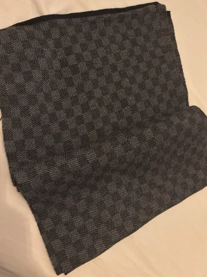 Gråsvart halsduk med rutmönster - Snygg halsduk i grått och svart med diskret rutmönster. Tunt vävt material som känns mjukt och följsamt. Perfekt accessoar för att lyfta din outfit med en stilren touch. Passar både till jacka och kappa.
