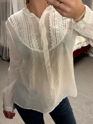 Vit blus med spetsdetaljer - Säljer en vit blus i tunn, luftig bomull med dekorativa spetsdetaljer och veck framtill. Blusen har ståkrage, knäppning upptill och långa ärmar.