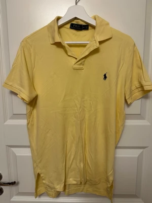 Pikétröja från Polo Ralph Lauren - Klassisk gul pikétröja från Polo Ralph Lauren med korta ärmar och den ikoniska mörkblå loggan broderad på bröstet. Tröjan har krage och knappar vid halsen. Perfekt för en fräsch och stilren look. Stor i storleken så passar S/M.