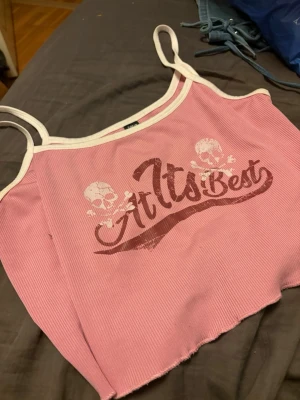 Rosa ribbad linnetopp med tryck - Trendig rosa linnetopp med vita kanter och smala axelband. Toppen har ett tryck med texten 'At Its Best' och två dödskallar. Ribbad struktur och croppad passform ger en cool vibe.