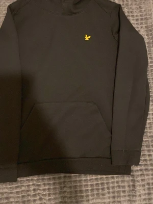 Svart hoodie från Lyle & Scott - Snygg svart hoodie från Lyle & Scott med klassisk gul logga på bröstet. Hoodien har huva med dragsko och en stor känguruficka framtill. Tillverkad i mjukt material som är skönt att bära. Perfekt för en avslappnad och stilren look.