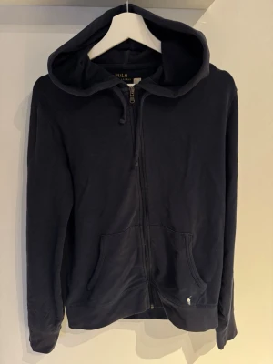 Mörkblå hoodie från Polo Ralph Lauren - Snygg mörkblå hoodie från Polo Ralph Lauren med dragkedja framtill och två fickor. Klassisk huva och liten vit logga broderad vid ena fickan. Tillverkad i mjukt bomullsmaterial som är skönt att ha på sig. Perfekt för en avslappnad och stilren look.