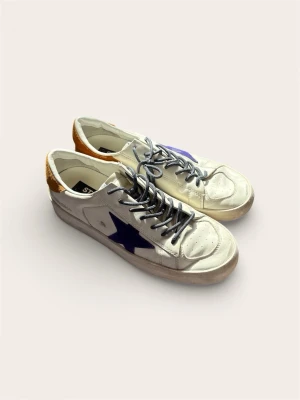 Golden Goose Stardan (Kvitto från NK) – Slutsåld modell - Säljer ett par exklusiva Golden Goose-sneakers i modellen "Stardan". En basket-inspirerad modell i vitt skinn med detaljer i lila (stjärnan) och hälkappa i orange mocka. Detta är en slutsåld färgställning som inte längre går att få tag på.  Skorna är inköpta på NK i Stockholm och originalkvitto medföljer vid köp.  Skick: 9/10 – Mycket fint skick! Endast använda ett fåtal gånger. Storlek: 41 (Sitter som 42-43). Originalbox finns ej, men levereras med kvitto.  Nypris: 6000 kr.