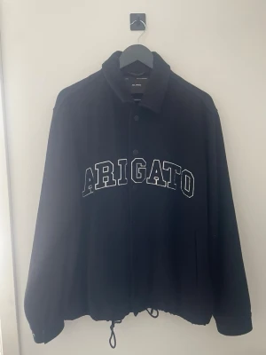 Svart jacka från Axel Arigato - Svart jacka från Axel Arigato med stor vit text på bröstet. Jackan har knappar framtill, snörning i nederkant och en klassisk krage. Jackan är i bra skick och har inga skador