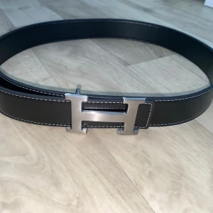Svart bälte med H-spänne  - Snyggt svart bälte med det ikoniska H-spännet i silverfärg. Bältet är gjort i slätt skinn med vita kontrastsömmar längs kanterna. Perfekt accessoar för att lyfta vilken outfit som helst.