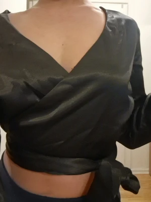 Svart omlottopp med knytband - Svart omlottblus med långa ärmar och knytband i midjan. Blusen har en v-ringning fram och en öppen rygg.