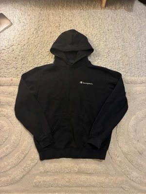 Svart hoodie från Champion - Säljer en svart hoodie från Champion med klassisk logga broderad på bröstet. Hoodien har en stor huva och magficka framtill. Tillverkad i mjukt bomullsmaterial och har ribbade muddar vid ärmslut och nederkant. Perfekt för en avslappnad och sportig stil.