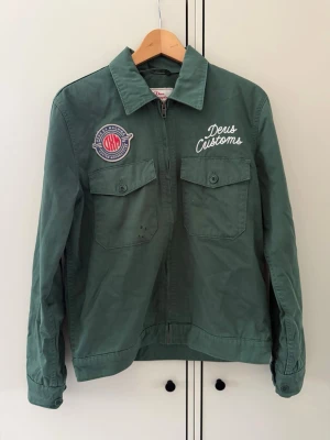 Grön overshirt från Deus ex machina - Snygg grön overshirt från Deus Customs med stora broderade patches på både bröstet och ryggen. Två bröstfickor med lock och knappar, klassisk krage och tryckknappar vid ärmslut. Materialet är kraftig bomull, perfekt för en streetwear-look.