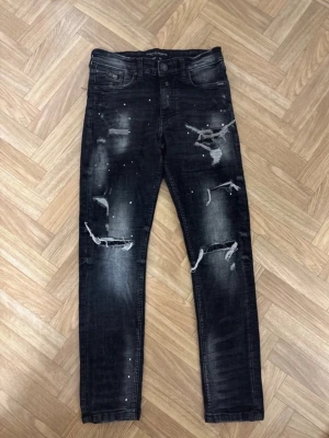 Svarta slitna skinny jeans Only & Sons - Säljer ett par svarta skinny jeans från Only & Sons med slitningar, hål och vita färgstänk på framsidan. Jeansen har fem fickor och en tvättad look som ger en cool och trendig vibe. Perfekta för dig som gillar streetstyle.