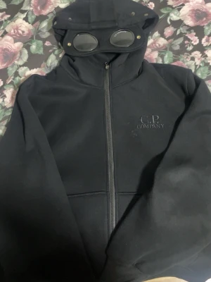 Svart hoodie med glasögon från C.P. Company - Säljer en svart hoodie från C.P. Company med ikoniska inbyggda glasögon i huvan. Dragkedja framtill och diskret logga på bröstet. Tillverkad i mjukt material som känns skönt mot huden. Perfekt för dig som vill sticka ut med en unik detalj i streetwear-stil.