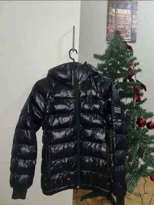 Svart dunjacka Peak Performance - Snygg svart dunjacka från Peak Performance med glansig finish och huva. Jackan har dragkedja framtill, två fickor och en tydlig logotyp på ärmen. Perfekt för kalla vinterdagar och håller dig varm med sin vaddering.