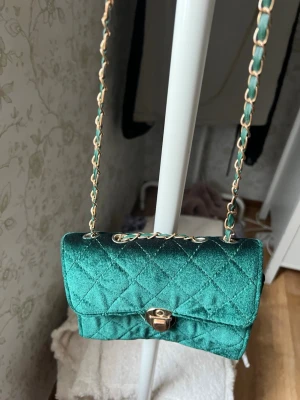 Grön quiltad axelväska med kedja - Snygg liten axelväska i smaragdgrönt sammetstyg med quiltat mönster. Väskan har guldfärgad kedjerem och ett vridlås framtill. Perfekt för att lyfta vilken outfit som helst med en poppig färg och trendig look.