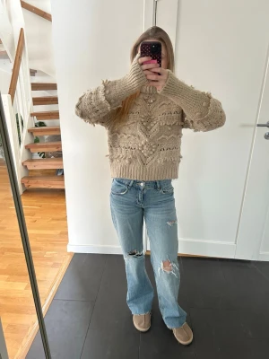 Beige stickad tröja med volangdetaljer - Säljer en mysig beige stickad tröja med volangdetaljer. Tröjan har långa ärmar och en hög krage, perfekt för kyligare dagar. Passar bra till både jeans och kjol.