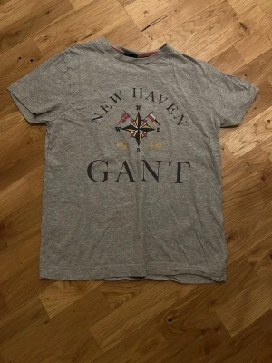 Grå T-shirt från GANT med tryck - Snygg grå T-shirt från GANT med stort tryck på bröstet: 'NEW HAVEN', kompassros och flaggor samt årtalet 1949. Klassisk rund hals och korta ärmar. Perfekt för en avslappnad och stilren look.
