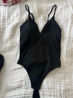 Svart body med smala axelband H&M - Snygg svart body från H&M med smala justerbara axelband och v-ringning. Tillverkad i mjukt stretchigt material med tryckknappar i grenen. Perfekt att styla till olika outfits för en trendig look.