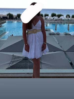 Vit kort klänning med broderi och bälte - Säljer en söt vit kort klänning med v-ringning, tunna axelband och volangkjol. Klänningen har broderade detaljer och ett brett beige bälte i midjan som ger en snygg siluett. Perfekt för varma dagar och sommarens alla tillfällen.