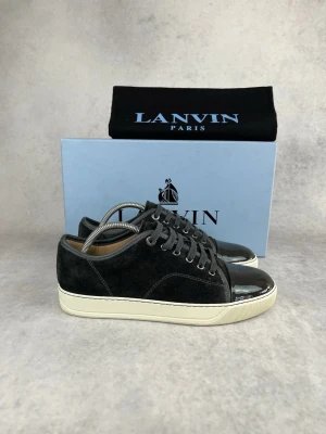 Svarta Lanvin sneakers med lackdetaljer - Snygga svarta sneakers från Lanvin med ovandel i mocka och glansiga lackdetaljer på tån. Skorna har vita sulor och klassisk snörning. Perfekta för dig som vill ha en stilren men ändå unik sneaker i garderoben.
