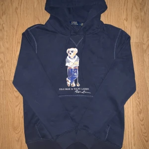 Mörkblå Polo Bear hoodie Ralph Lauren - Säljer en mörkblå hoodie från Ralph Lauren med Polo Bear-tryck på bröstet. Tröjan har huva med snörning, ribbade muddar och känguruficka. Materialet är mjuk bomull och polyesterblandning, perfekt för chill dagar. Klassisk streetstil med snyggt motiv.