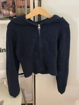 Mörkblå hoodie från Gina Tricot - Mysig mörkblå hoodie från Gina Tricot med dragkedja framtill och huva. Tröjan är ribbstickad nedtill och vid ärmsluten, samt har en croppad passform. Perfekt för en avslappnad och trendig look.