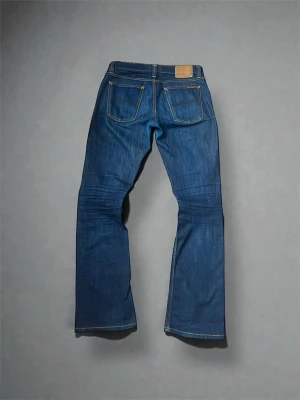 Nudie bootcut jeans - Säljer ett par klassiska blå jeans från Nudie Jeans med raka ben och femficksmodell. Jeansen har kontrastsömmar och en patch i läderimitation bak i midjan. Tillverkade i slitstark denim av bomull. Perfekta för en avslappnad och stilren look.