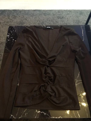 Svart topp med knutdetaljer från bikbok  - Trendig svart långärmad topp från bikbok med djup V-ringning och tre snygga knutdetaljer framtill. Tillverkad i ett ribbat, stretchigt material som sitter snyggt på kroppen. Perfekt för dig som gillar unika detaljer och vill sticka ut.