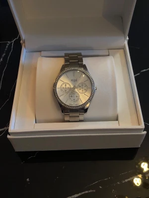 Silverfärgad klocka från Regal - Stilren klocka från Regal i silverfärgat metallarmband och boett. Urtavlan är ljus med tre mindre visartavlor och stenar runt boetten för extra bling. Perfekt accessoar för att lyfta din outfit. Batteriet behövs bytas, om man vill använda klockan som en accessoar kan man skippa att byta batteri. Nypris ca 399. Priset kan diskuteras! Boxen följer ej med. 