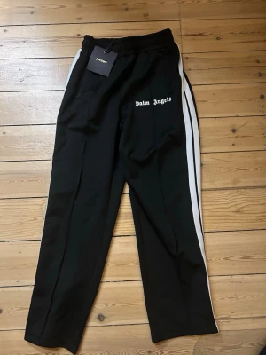 Palm Angels svarta mjukisbyxor - Svarta mjukisbyxor från Palm Angels med vita sidoränder och logga tryckt på vänster sida. Byxorna har elastisk midja och raka ben för en chill och loose vibe. Perfekta för dig som vill ha streetwear med stil.