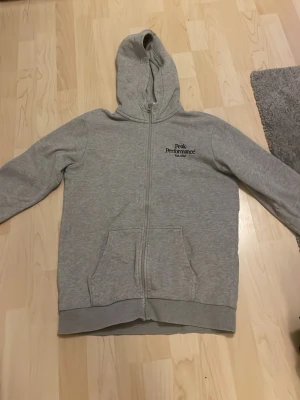 Grå hoodie från Peak Performance - Snygg grå hoodie från Peak Performance med dragkedja framtill och två stora fickor. Klassisk huva och diskret logga på bröstet. Tillverkad i mjukt bomullsmaterial som är skönt att ha på sig. Perfekt för en avslappnad och sportig stil.