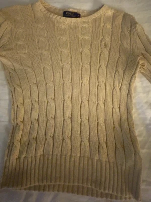 Beige kabelstickad tröja Polo Ralph Lauren - Klassisk beige kabelstickad tröja från Polo Ralph Lauren. Tröjan har rund halsringning, långa ärmar och ribbade muddar. Tillverkad i mjukt material som ger en skön passform och tidlös look. Perfekt till jeans eller kjol för en clean stil.