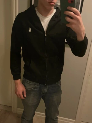 Svart hoodie från Polo Ralph Lauren - Svart hoodie med dragkedja från Polo Ralph Lauren. Klassisk design med vit broderad logga på bröstet och två fickor framtill. Tillverkad i mjukt material och har en skön huva. Perfekt för en avslappnad och stilren look.