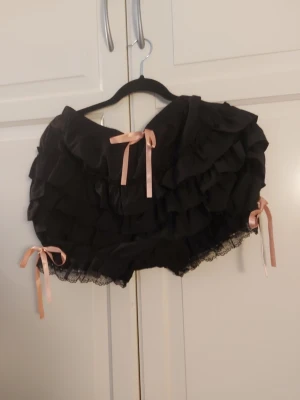 Svarta volangshorts med rosetter - Supersöta svarta pyjamasshorts med flera lager volanger och spetskant. Dekorerade med ljusrosa satinrosetter framtill och på sidorna för extra gullig look. Perfekta för mysiga hemmakvällar eller när du vill känna dig extra fin i loungewear.